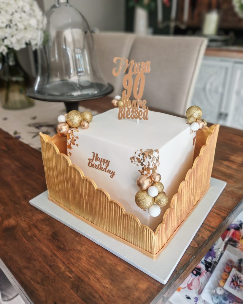 Square white and gold 90th birthday cake with rose-gold spheres, gold leaf and a Years Blessed topper
